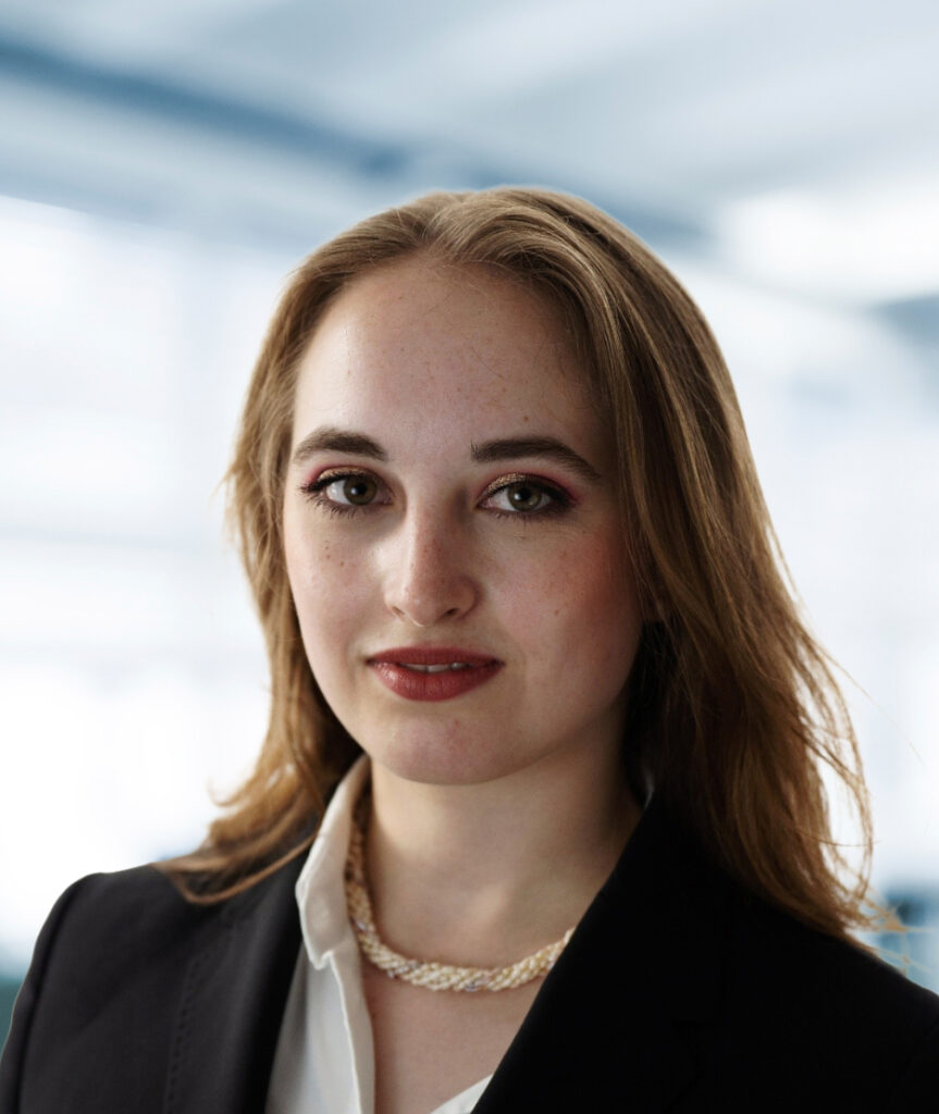 Das Bild zeigt Lisa Kukuk. Sie trägt ein dunkles Jackett über einer hellen Bluse sowie eine helle Halskette, der Hintergrund ist hell und unscharf gehalten, wodurch die Person deutlich im Vordergrund steht.