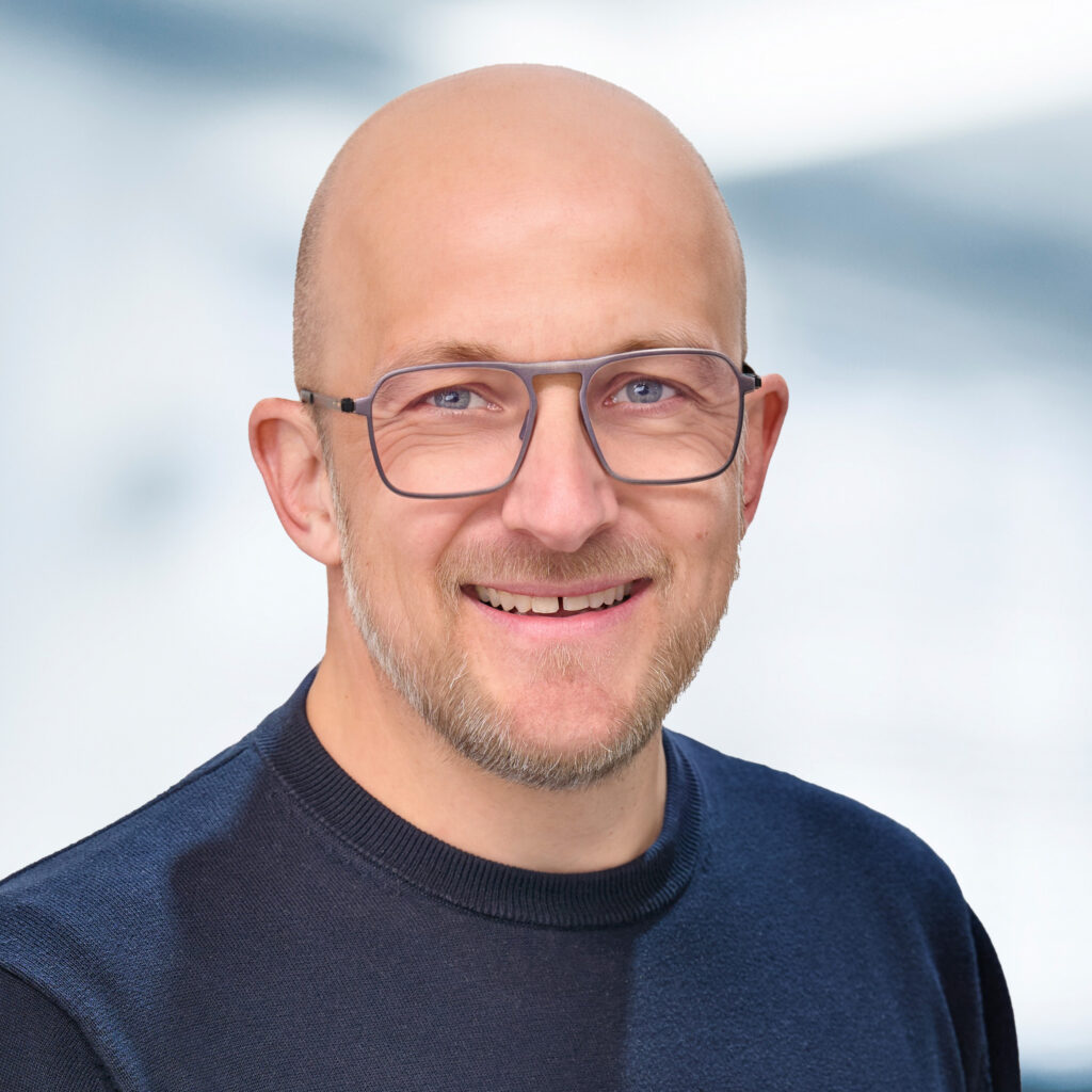 Das Bild zeigt Alexander Dütz. Er trägt einen dunklen Pullover und eine Brille, der Hintergrund ist hell und unscharf gehalten, wodurch die Person deutlich im Vordergrund steht.